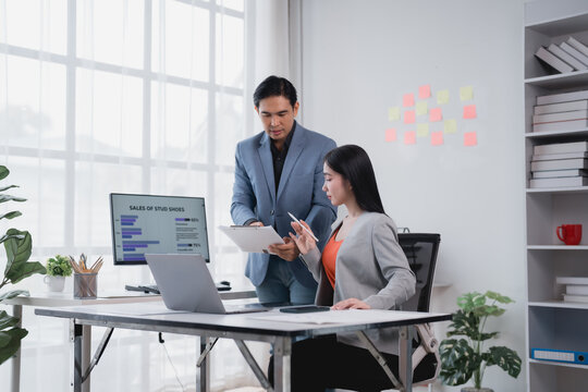 Two asian businesspeople are discussing sales data in a modern office, analyzing charts and reports on a computer screen and clipboard, collaborating on a project and sharing insights