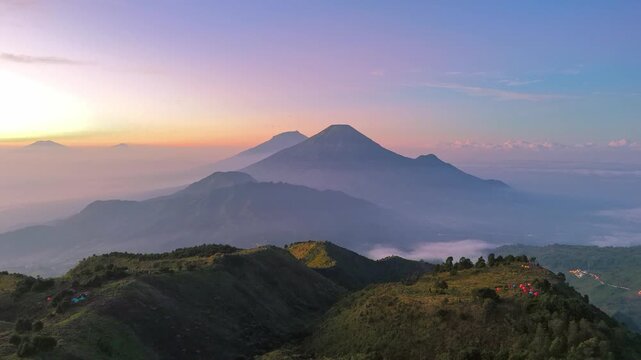 Aerial view of lush greenery mountain ridge at sunrise with dramatic sky and volcanic peaks in the distance. Amazing sunrise scenery of Mount Prau, Indonesia.