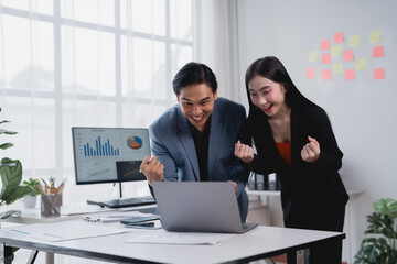 Celebrating success, two Asian businesspeople smile and raise their fists while looking at a laptop in a modern office, embodying teamwork and achievement