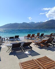 Scenic vertical view of Tivat beach with wooden sunbeds by calm turquoise sea, framed by lush mountains and bright sky with white clouds.