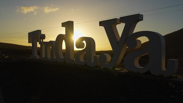 Sculpture with the word "Tindaya" in Fuerteventura at sunset, sunlight rays passing through metal letters, mountains in background, cinematic camera movement