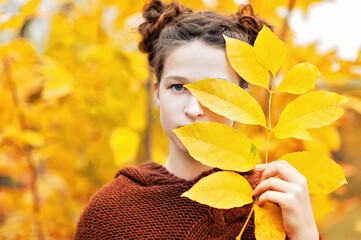 Autumn outdoor portrait with vibrant foliage. Girl wearing a brown sweater holding vibrant yellow leaves in front of her face with an autumn forest background