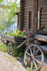 Obraz premium Beautiful garden composition with a wooden cart, flowers, and a wheel in front of a log house. Element of rustic style and coziness.