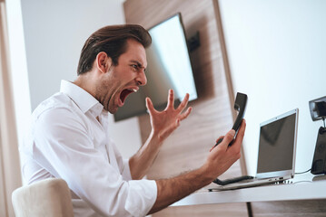 A young man wearing a white shirt is sitting at a desk, yelling into his phone while surrounded by...