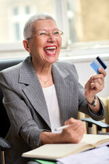 A cheerful elderly woman with short gray hair is excitedly reviewing her finances while holding a...