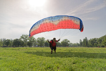 A skydiver approaches the ground while maneuvering a colorful parachute in a grassy park. Friends...