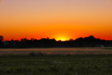 A peaceful rural landscape where the setting sun hides behind the silhouettes of the forest on the horizon. The warm orange-red sky creates a cozy atmosphere and a sense of calm.