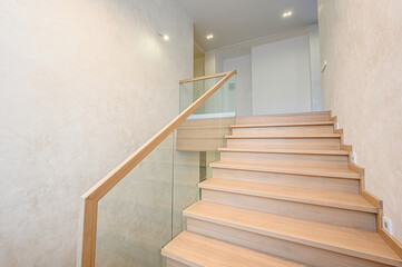bright indoor scene features wooden stairs with a glass railing. Walls are light beige. The space...