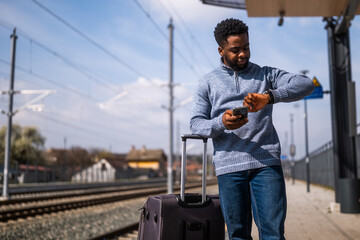 African American commuter stressed about travel delays, looking at his clock while standing on...