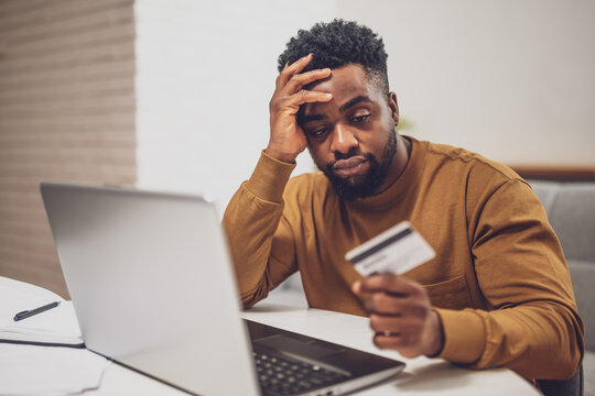 African man feeling stressed while using laptop and credit card at home. He is dealing with financial problems.