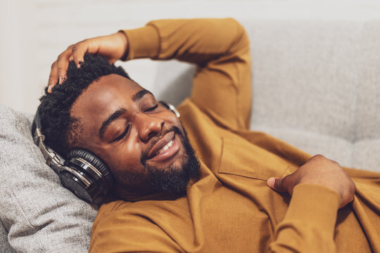 Portrait of happy young African American man enjoying listening to music on headphones and relaxing on sofa at his home.