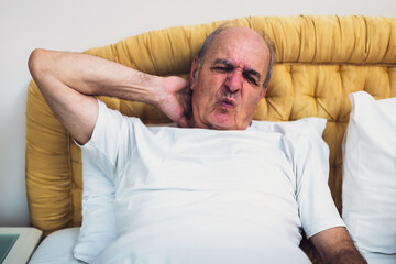 Senior man lying in bed, holding his neck in pain after waking up.