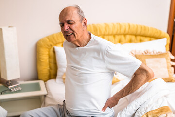Senior man holding his back in pain while sitting on bed in the morning.