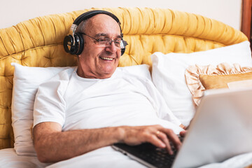 Smiling senior man in pajamas using laptop and wearing headphones while sitting in bed.	