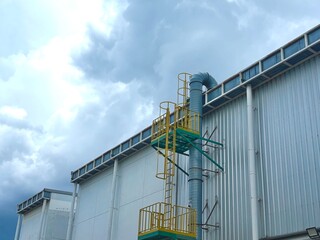 Fototapeta premium A large industrial building with a ventilation system and a maintenance ladder against a dramatic, cloudy sky. This image represents modern factory infrastructure and environmental control.