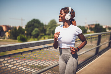Muscular African - American woman with headphones on jogging outdoors on urban city bridge.