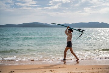 Surfer walks along the shoreline carrying a surfboard in the early morning light near a tranquil...