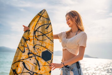 Young woman examines yellow surfboard while standing on sunny beach during late afternoon
