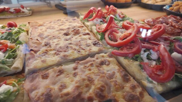 Italian pizza al taglio with slices topped with fresh arugula, cherry tomatoes, smoked salmon, and mozzarella in bakery display, showcasing traditional street food