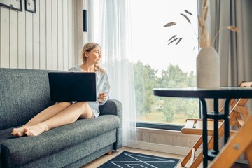 woman on sofa with laptop gazing thoughtfully out large window in bright room with wooden walls,...