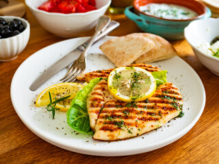 Greek style food: grilled chicken breast with lemon, pita bread,  tzatziki sauce on wooden table	