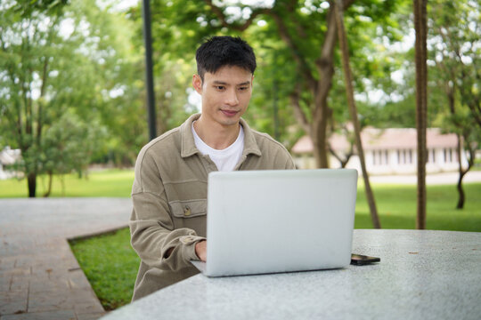 Smiling asian man working remotely on a laptop in a park.