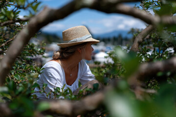 A serene shot of a woman peeking through lush greenery while wearing a summer hat captures the essence of nature and relaxation during a sunny day outdoors.
