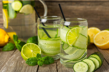 Cucumber water with lemon, lime and mint on a wooden table. Water infused with sliced ​​cucumber in a drinking glass. Tonic refreshing drink. Cucumber lemonade.Summer natural cooling drink.Copy space