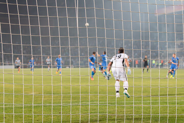 Focused on the goal net during a soccer match. Blurred view of the goalkeeper and players on the green pitch, capturing the action and atmosphere of a live game.