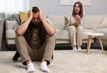 Betrayal. Angry woman with smartphone shouting at her offended husband on sofa at home, selective...
