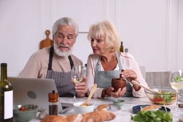Elderly couple with laptop cooking together and drinking white wine at table indoors
