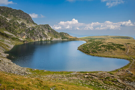 A beautiful glacial lake, part of the Seven Rila Lakes, sits in a rocky mountain cirque with a hiking trail along its shore under a blue, cloudy sky, Kidney Lake, Rila Mountain, Bulgaria.