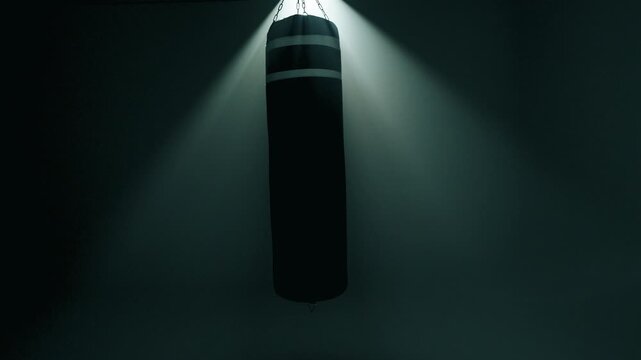 A black boxing bag gently swings left to right in a dark, smoky gym. Intense backlight shines from behind, creating a glowing halo effect. Scene evokes anticipation or reflection around training