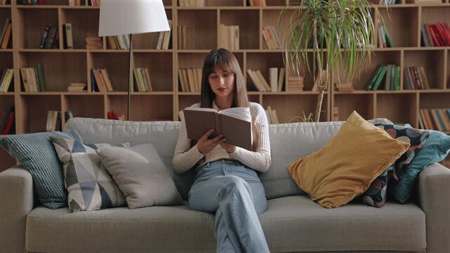 Charming Caucasian woman crossing legs on sofa and holding book while gazing upward thoughtfully. Reflecting on passage or imagining story scene in peaceful room surrounded by pillows and books.