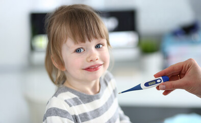 Smiling child sits indoors with a thermometer in hand while showing signs of playful curiosity during a cozy morning