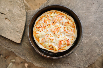A close-up shot of a Hawaiian pizza in a black pan on a rustic wooden surface.