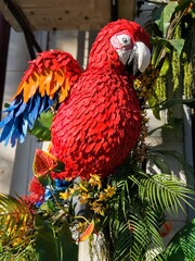 Scarlet macaw intricately designed with vibrant red, blue, and orange feathers perched among lush green foliage and tropical flowers, showcasing a lifelike yet artistic display.
