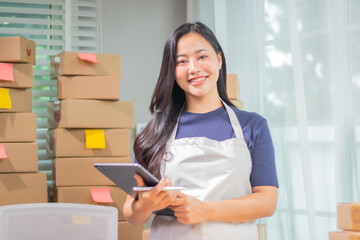 Asian entrepreneur checking shipping list on laptop while packing boxes in warehouse for his small...