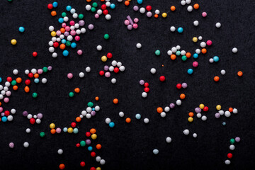 Multicolored round candy sprinkles scattered randomly on a dark textured background, showing a festive and playful food decoration concept