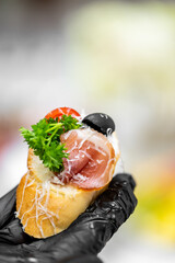 Close-up of an elegant canapé held in a black-gloved hand, featuring bread with prosciutto,...