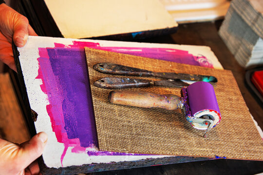 Hands of artist holding a textured printing plate with a purple inked brayer and palette knives arranged on top. Printmaking or monotype printing tools
