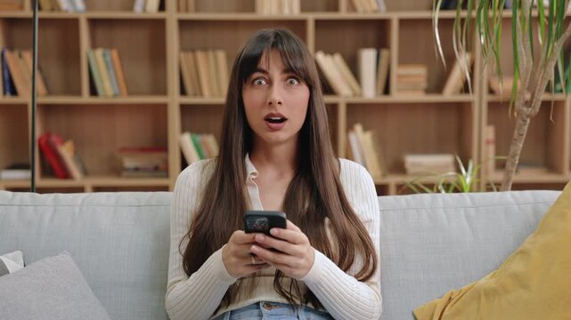 Caucasian woman with long dark hair reacting shocked while holding smartphone, sitting on couch in bright room. Surprised female looking at phone screen, leaning forward with widened eyes.