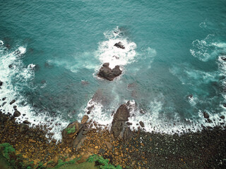 Aerial view of turquoise ocean waves crashing against rocky coastline and green vegetation.