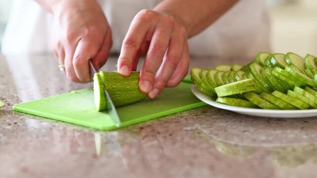 Close-up of female hands slicing fresh zucchini into thin pieces on a cutting board while preparing ingredients for homemade ratatouille in a bright kitchen setting
