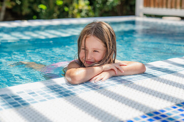 Cute smiling girl in a summer pool with blue refreshing water at an open-air resort. Bright sunny...