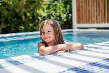 Cute smiling girl in a summer pool with blue refreshing water at an open-air resort. Bright sunny...
