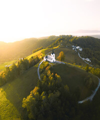 Drone photo of a small white church on a sunlit hilltop surrounded by autumn trees and countryside...