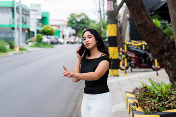 Confident Asian woman in black top and white pants on a phone call while gesturing or calling a taxi or car by the sidewalk on a city street setting.