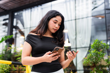 Asian woman in black off-shoulder top entering card details on phone, seemingly processing an online transaction outside a modern cafe setting.