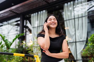 Fototapeta premium Beautiful Asian woman in a black off-shoulder top, joyfully smiling while talking on the phone outside a modern cafe setting.